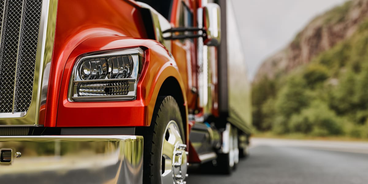 Close-up of a red semi-truck’s headlight, wheel, and chrome bumper traveling on a mountain highway, illustrating the causes of truck accidents.